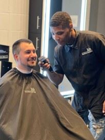 A barber using clippers on a seated man in a barbershop.