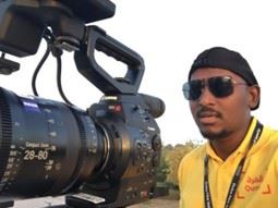 A man in a yellow shirt and cap operating a professional video camera outdoors.